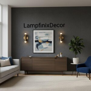 May include: A contemporary living room scene featuring a grey sofa, a dark wood cabinet, and a blue armchair. The wall displays a framed abstract painting and two gold sconces. The text "LampfinixDecor" is visible on the wall.