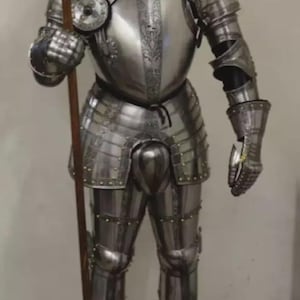 May include: Full suit of polished steel armor, including a helmet, breastplate, and leg and arm protection. The armor is highly detailed with decorative elements and rivets. A wooden spear is held in the right hand.