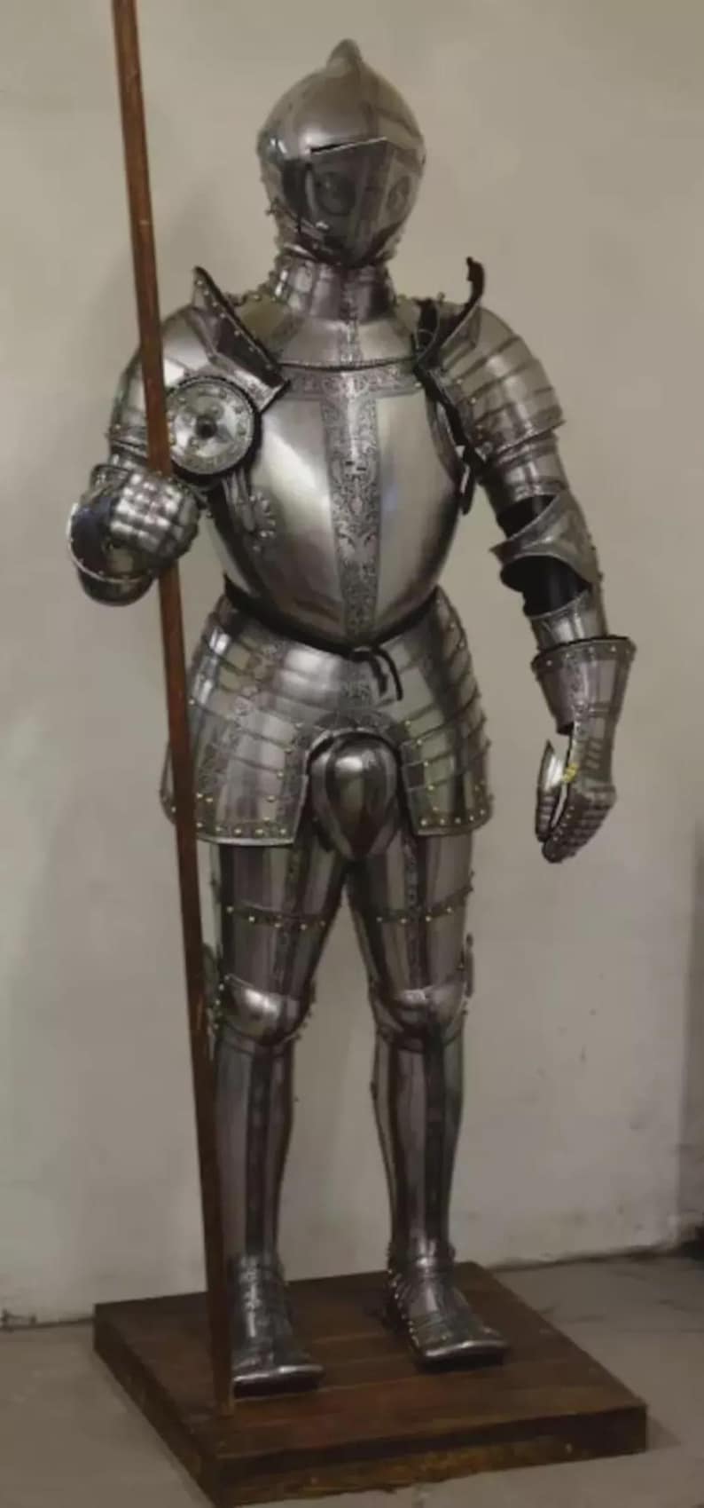 May include: Full suit of polished steel armor, including a helmet, breastplate, and leg and arm protection. The armor is highly detailed with decorative elements and rivets. A wooden spear is held in the right hand.
