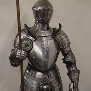 May include: A full suit of polished steel armor, including a helmet, chest plate, and leg protection. The armor is intricately detailed with decorative patterns and rivets. A wooden spear is held in the right hand. The armor is designed to protect the wearer.