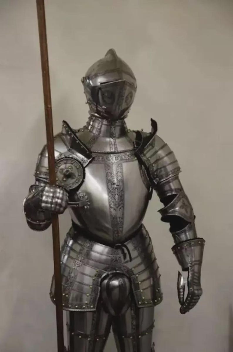 May include: A full suit of polished steel armor, including a helmet, chest plate, and leg protection. The armor is intricately detailed with decorative patterns and rivets. A wooden spear is held in the right hand. The armor is designed to protect the wearer.