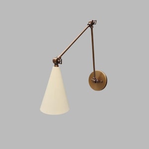 Op de afbeelding: Een bronzen wandlamp met een verstelbare arm en een conische, crèmekleurige lampenkap. De lamp heeft een ronde basis en scharnieren voor flexibele positionering, ideaal voor lees- of taakverlichting.