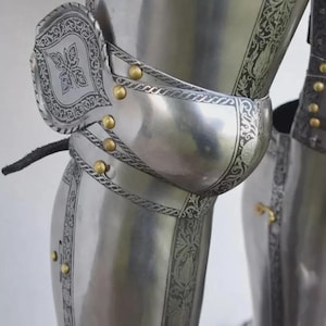 May include: Close-up of a section of medieval armor. The polished steel armor features intricate etched floral designs and is accented with gold-colored rivets. The image highlights the knee and leg protection, showcasing the craftsmanship of the historical garment.