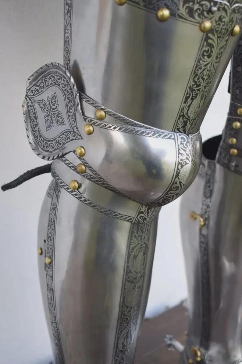 May include: Close-up of a section of medieval armor. The polished steel armor features intricate etched floral designs and is accented with gold-colored rivets. The image highlights the knee and leg protection, showcasing the craftsmanship of the historical garment.