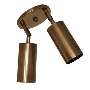 Puede incluir: Un foco de techo de dos luces de color latón. El accesorio tiene una base circular y dos cabezales de luz cilíndricos que se pueden ajustar. Los cabezales de luz están en ángulo y el accesorio está diseñado para uso en interiores.