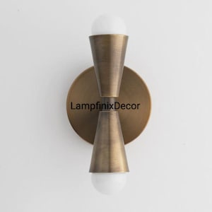 May include: A brass wall sconce featuring two conical shades, each housing a white, spherical bulb. The fixture has a circular backplate with the text "LampfinixDecor". It is mounted on a white wall. The sconce is a decorative lighting fixture.