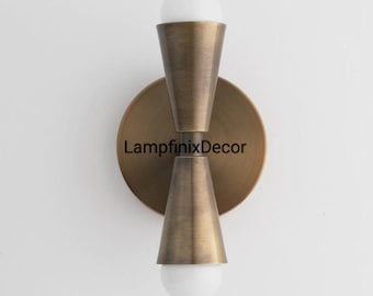 Pode incluir: Arandela de parede em tom de latão com duas cúpulas cônicas e uma placa traseira redonda. Cada cúpula contém uma lâmpada esférica branca. O texto "LampfinixDecor" está impresso na placa traseira. Montado em uma parede branca.