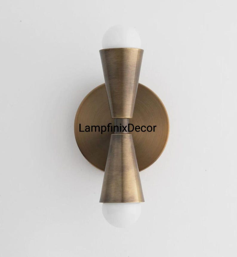 May include: A brass wall sconce featuring two conical shades, each housing a white, spherical bulb. The fixture has a circular backplate with the text "LampfinixDecor". It is mounted on a white wall. The sconce is a decorative lighting fixture.