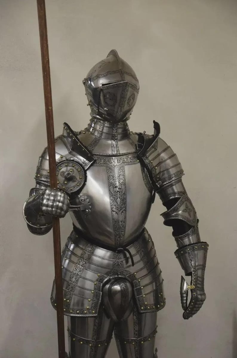 May include: A full suit of polished steel armor, complete with a helmet, chest plate, and articulated limbs. The armor is adorned with decorative etchings and rivets. A wooden spear is held in the right hand, suggesting a medieval or historical theme.