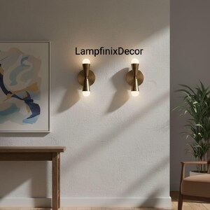 May include: Two brass wall sconces with a contemporary design, each with a circular base and an hourglass-shaped central section holding a spherical light bulb. The sconces are mounted on a textured white wall. The text "LampfinixDecor" is visible.