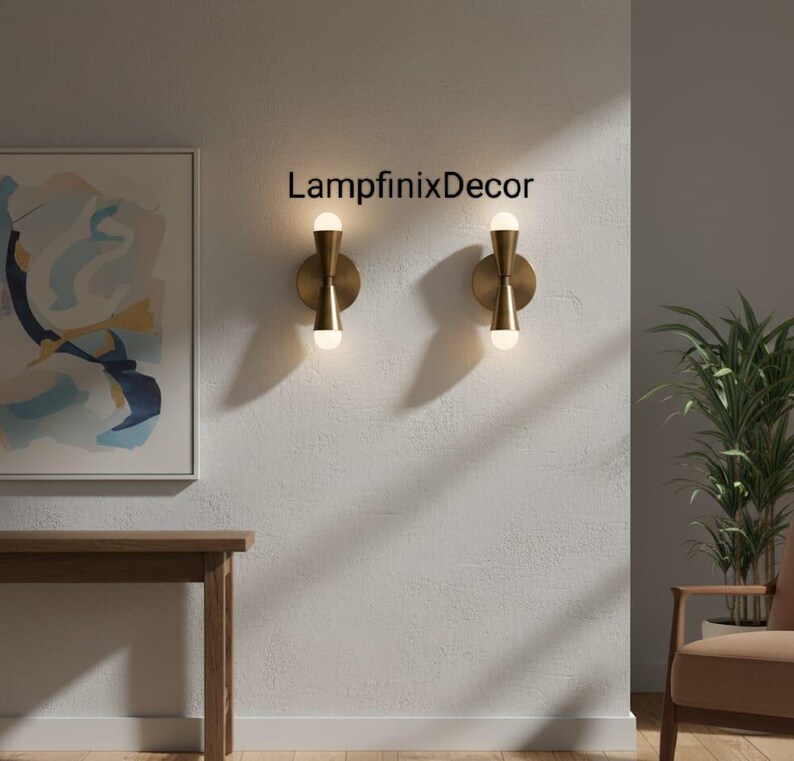 May include: Two brass wall sconces with a contemporary design, each with a circular base and an hourglass-shaped central section holding a spherical light bulb. The sconces are mounted on a textured white wall. The text "LampfinixDecor" is visible.