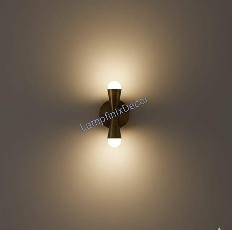 May include: A contemporary wall light with a brass finish, featuring two spherical light bulbs. The fixture has a distinctive hourglass shape and is mounted on a circular base. The text "LampfinixDecor" is visible on the image.