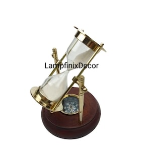 May include: An hourglass with a brass frame and a wooden base. The hourglass contains white sand and is mounted on a wooden base that also features a compass. The text "LampfinixDecor" is visible on the image.