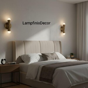 May include: A bedroom scene featuring two brass wall sconces with glowing bulbs. The beige headboard and white bedding are visible. A brown throw and the text "LampfinixDecor" on the wall complete the scene.