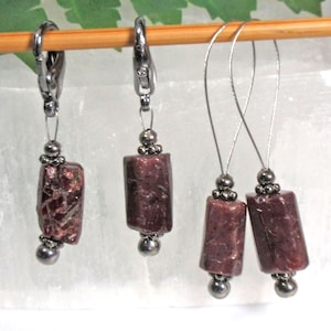 May include: Three pairs of earrings with dark red rectangular beads and silver-colored accents. Two pairs have a wire hook, and one pair has a hinged clasp. The earrings are displayed on a wooden dowel against a white background.