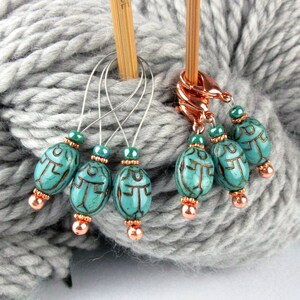Peut inclure: Six marqueurs de points en forme de scarabée turquoise bleu avec des perles en cuivre et des boucles en fil d'argent. Les marqueurs sont sur une pelote de laine grise avec deux aiguilles à tricoter en bois.