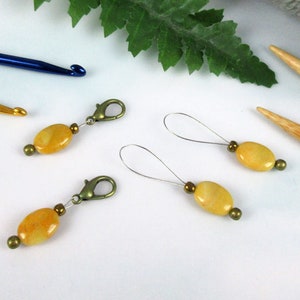 May include: Three yellow stone stitch markers with metal clasps and two yellow stone stitch markers with metal loops. The stitch markers are on a white surface.