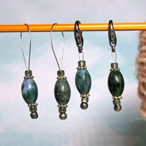 May include: Four stitch markers hanging from a yellow knitting needle. Two markers feature silver wire loops, while the other two have dark metal lobster clasps. Each marker includes a green moss agate bead, small silver spacers, and dark beads.