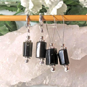 May include: Four dark gray, rectangular gemstone stitch markers with silver-colored accents hang from a wooden knitting needle. The markers have a polished, reflective surface and are suspended by thin silver wires.