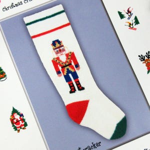 May include: A white knit Christmas stocking featuring a central nutcracker figure in red, blue, and gold. The stocking has a green and red striped cuff at the top, a red heel, and a green toe. The design is knitted into the white fabric.