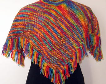 May include: A knitted poncho featuring a vibrant, multi-coloured rainbow yarn pattern with a high neck collar. The garment is constructed with a textured stitch and finished with long, loose fringe tassels along the entire bottom hemline.
