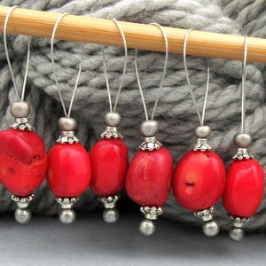 May include: Seven red coral stitch markers with silver metal caps and beads. The markers are hanging from a wooden knitting needle.
