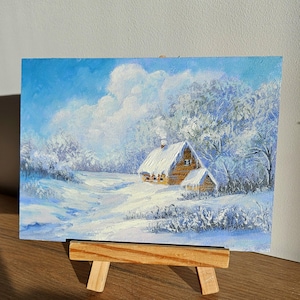 May include: Oil painting depicting a snow-covered cabin in a winter scene. The artwork shows a wooden cabin with a snow-covered roof, surrounded by snow-laden trees and a bright blue sky. The painting is displayed on a small wooden easel.