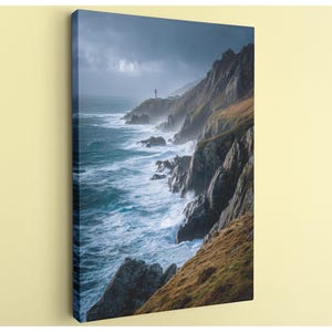 May include: A canvas print of a coastal scene featuring a lighthouse on a rocky cliff. The ocean waves crash against the rocks, creating a dramatic display of white foam. The sky is overcast, adding to the moody atmosphere. The landscape is a mix of brown, green, and gray.