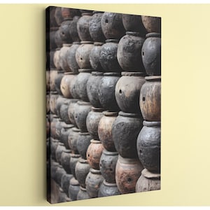 May include: A canvas print featuring a wall of stacked, weathered, dark brown and gray ceramic pots. Each pot has a rounded shape with a small opening. The pots are arranged in a grid pattern, creating a textured, rustic aesthetic.