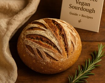 May include: A round loaf of sourdough bread with decorative cuts on top, next to a sprig of rosemary and a book titled "Gluten-Free Vegan Sourdough Guide + Recipes". The bread is golden brown with a light dusting of flour.