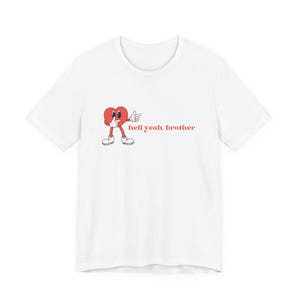 May include: White t-shirt featuring a cartoon heart character with arms, legs, and thumbs up gesture. The text "hell yeah, brother" is printed in red. The design is centered on the front of the shirt.