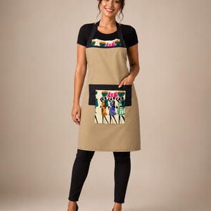 May include: A tan apron with a black pocket and trim, featuring a colorful illustration of women carrying baskets on their heads. The apron is worn over a black top and pants.
