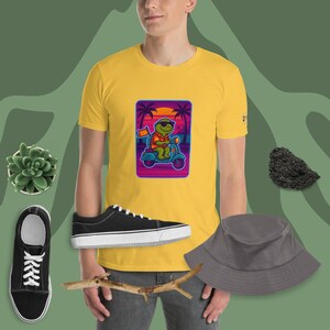 Op de afbeelding: Geel T-shirt met een cartoon schildpad op een scooter tegen een zonsondergang. De afbeelding bevat zwarte sneakers, een grijze vissershoed en een vetplant. De stijl is casual en leuk.