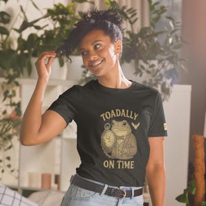 Könnte beinhalten: Schwarzes T-Shirt mit einer Cartoon-Frosch-Illustration, die eine Taschenuhr hält und einen Anzug trägt, mit dem Text "TOADALLY ON TIME". Das Design ist in Braun- und Goldtönen gehalten. Das Shirt hat einen Rundhalsausschnitt.