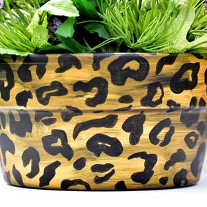 May include: A decorative flower pot with a gold base and black leopard print design. The pot is filled with green foliage and purple flowers, creating a vibrant contrast. The pot is suitable for indoor or outdoor use.