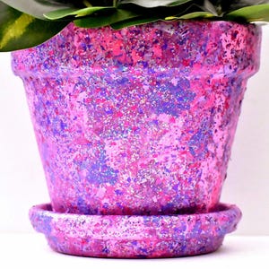 May include: A tapered plant pot with a matching saucer, featuring a textured finish in vibrant shades of pink, purple, and blue with a heavy application of silver glitter throughout the surface, containing a green leafy plant at the top.