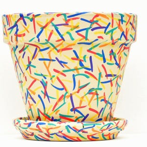 May include: A cream-colored ceramic plant pot with a matching saucer, featuring a hand-painted pattern of short, scattered brushstrokes in vibrant shades of red, blue, yellow, orange, and green across the entire exterior surface.