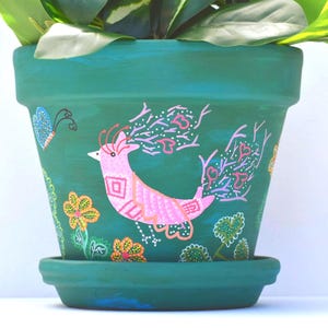May include: A teal-colored ceramic flower pot with a matching saucer. The pot is hand-painted with a pink bird, flowers, and a butterfly. The pot is filled with green leafy plants.