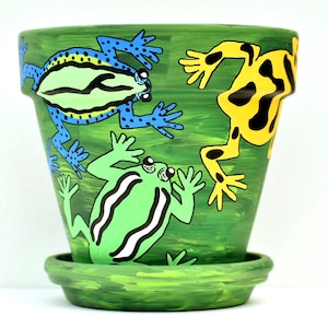 May include: A green terracotta plant pot with a matching saucer, hand-painted with a vibrant scene featuring three frogs. One frog is blue with black spots, one is yellow with black spots, and one is light green with white and black stripes.