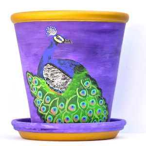 May include: A tapered terracotta plant pot with a matching saucer, featuring a hand-painted purple background and a detailed peacock illustration. The peacock displays vibrant blue, green, and gold plumage. The rim and base are painted in yellow.