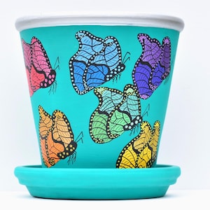 May include: A turquoise ceramic flower pot with a matching saucer. The pot is decorated with colorful butterfly illustrations in shades of pink, blue, green, orange, and yellow. The rim of the pot is white.