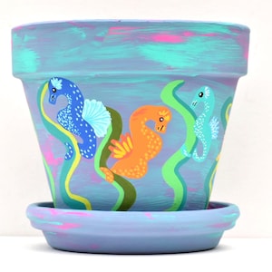 May include: A hand-painted terracotta flower pot and matching saucer featuring a light purple base with brushstrokes of turquoise and pink. The exterior is decorated with colorful seahorses in blue, orange, and light green, surrounded by wavy green seaweed.