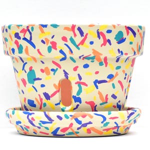 May include: A cream-colored ceramic flower pot with a matching saucer, both decorated with colorful abstract brushstrokes in blue, yellow, orange, pink, and teal. The pot has a drainage hole and is suitable for planting.