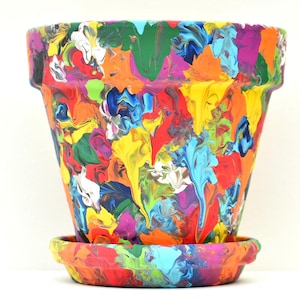 May include: A tapered plant pot with a matching saucer, both featuring a vibrant, multi-colored paint drip design. The surface is covered in swirling patterns of bright yellow, orange, red, blue, green, purple, and white paint in a thick texture.