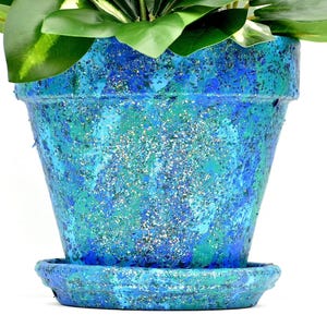 May include: A tapered flower pot with a matching saucer, featuring a textured finish in shades of blue, teal, and green with silver glitter accents. The pot contains a green plant with broad leaves extending from the top opening.