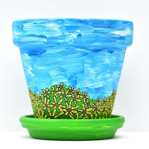 May include: A hand-painted ceramic flower pot with a blue sky design and a green base. The pot features a painted landscape with yellow flowers at the bottom. The pot sits on a matching green saucer.