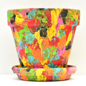 May include: A tapered plant pot with a matching saucer, featuring a vibrant, multi-colored abstract paint drip design. The surface is covered in swirling patterns of neon pink, bright yellow, orange, lime green, red, black, and turquoise paint.