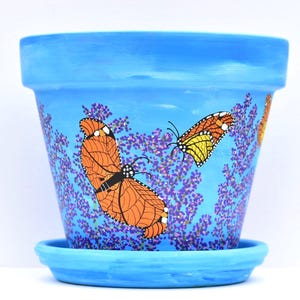 May include: A hand-painted, light blue ceramic flower pot with a matching saucer. The pot is decorated with orange and yellow butterflies and purple floral accents. The pot is designed for indoor or outdoor use.