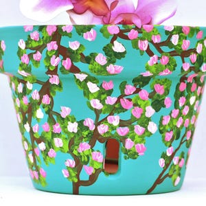 May include: A hand-painted turquoise ceramic flower pot. The pot is decorated with a floral design featuring brown branches, green leaves, and pink and white blossoms. The pot has a rectangular drainage hole.