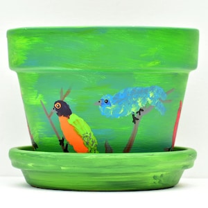 May include: A bright green terracotta plant pot with a matching saucer, hand-painted with a scene featuring a blue bird perched on a brown branch and a second bird with a black head and orange chest sitting on a separate brown branch.